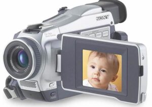 A silver Sony DCR-TRV18 MiniDV Handycam camcorder shown from a three-quarter front angle, showcasing its optical zoom lens, built-in stereo microphone, and a side-mounted flip-out LCD screen showing a blue display.